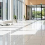 spotless, highly reflective polished light-colored tile floor in a modern, sunlit office lobby with full-height windows and wood paneling.